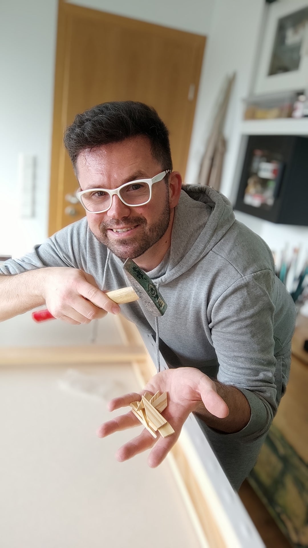 Der Künstler blickt lächelnd in die Kamera, während er mit einem Hammer kleine Holzkeile in den Keilrahmen einer großen Leinwand schlägt.