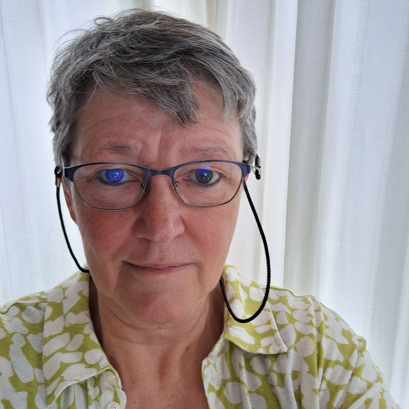 Porträtfoto einer Frau mit Brille, grauem Kurzhaarschnitt und gemusterter Bluse, vor hellem Vorhang.