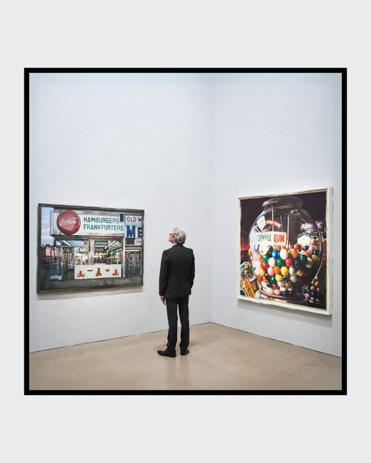 Das fotorealistische Digitalgemälde "Berlin" aus der Serie "Rueckenfiguren" von nostrokes zeigt einen Mann in einem schwarzen Anzug, der von hinten in einer Kunstgalerie steht. Er betrachtet zwei große, farbige Gemälde im Stil des Fotorealismus an einer weißen Wand. Das quadratische Werk ist in einer limitierten Edition auf Leinwand gedruckt und in der Online-Galerie ARTSPACER erhältlich.