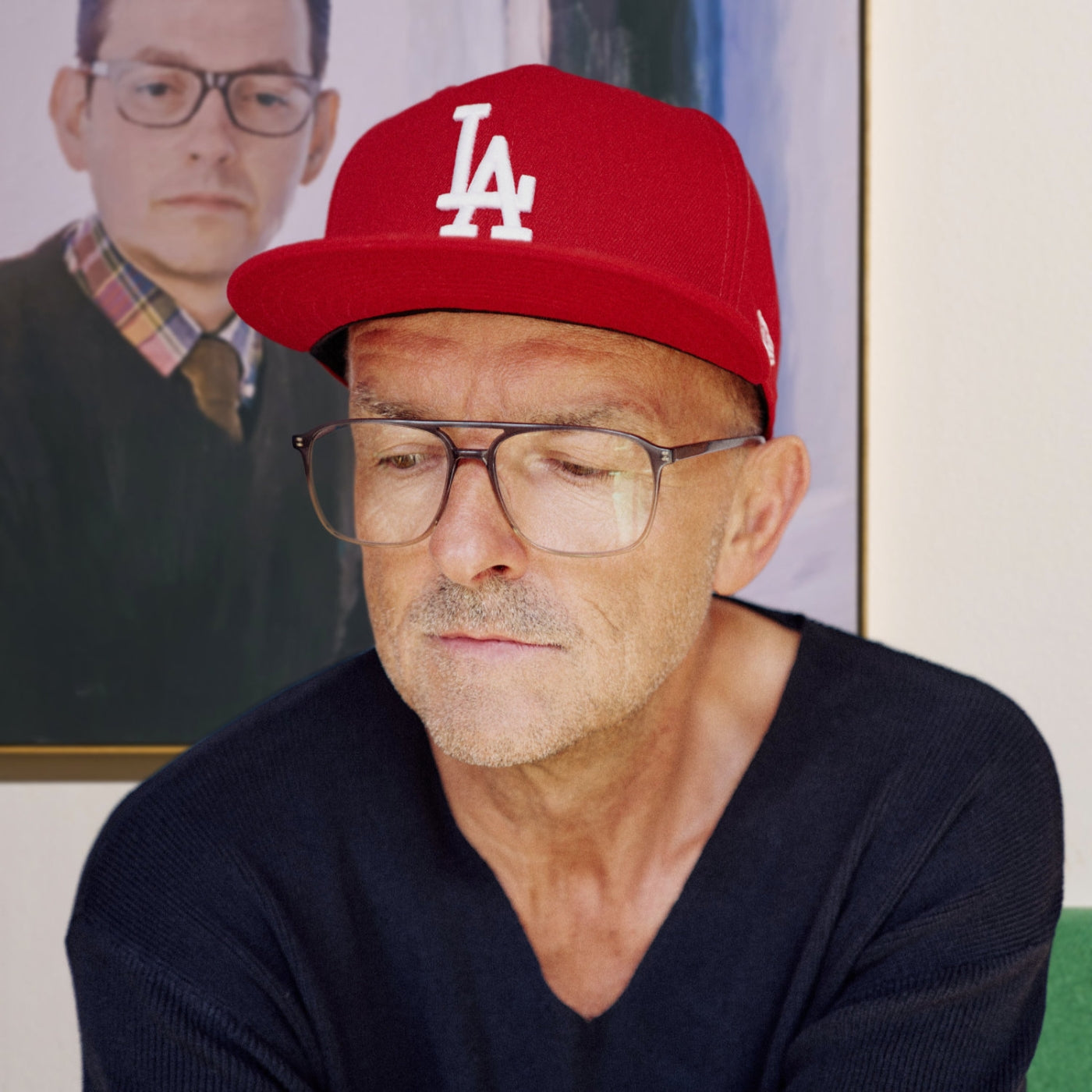 Profilbild des Künstlers nostrokes für die Online-Galerie ARTSPACER. Das Bild zeigt ihn in entspannter Pose mit roter Mütze, während im Hintergrund eines seiner typischen, malerischen Porträts auf Leinwand zu sehen ist.