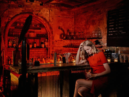 Das fotorealistische Digitalgemälde "Chablis bei Schlamassel" von nostrokesbitte zeigt eine blonde Frau im roten Kleid, die melancholisch an einer rot beleuchteten Bar sitzt und ein Glas Wein hält. Das Werk ist in limitierter Auflage bei ARTSPACER erhältlich.