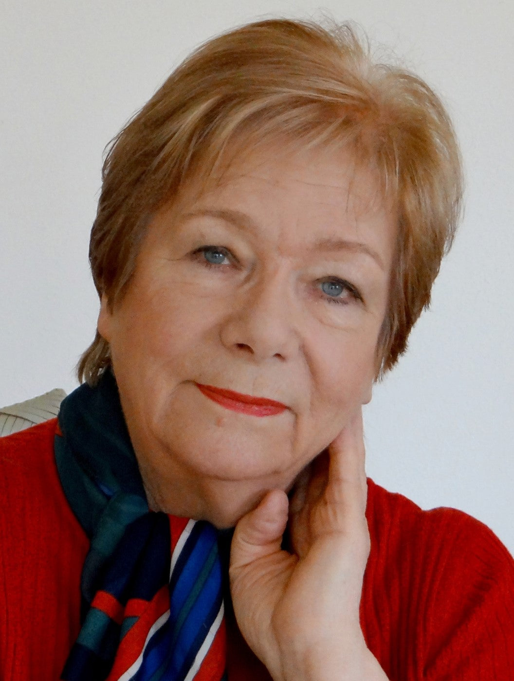 Porträt von Anke Huober, einer Künstlerin der Online-Galerie ARTSPACER. Sie ist eine Frau mittleren Alters mit kurzen blonden Haaren und trägt einen roten Pullover und einen blau-grün gemusterten Schal. Sie stützt ihr Kinn auf ihrer rechten Hand und lächelt leicht. 