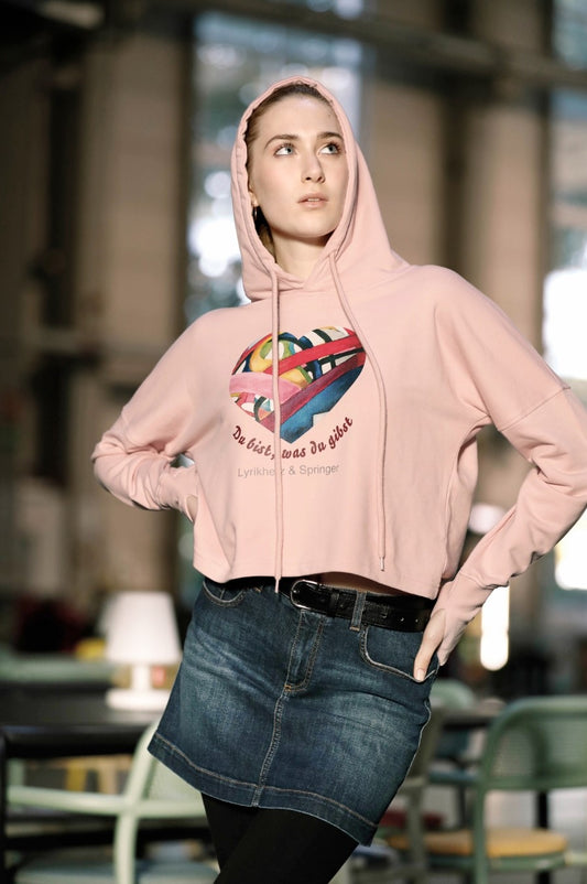 Ein kurz geschnittener, rosafarbener Hoodie mit überschnittenen Schultern. Extralange Bündchen mit Daumenloch und eine bedruckte Kunstgrafik auf der Vorderseite. Mode von Soulfashion Lyrikherz & Springer.