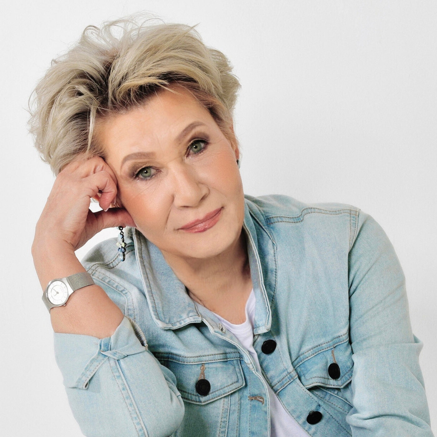 Ein Porträtfoto der Künstlerin Gabriele Springer vor einem hellen, neutralen Hintergrund. Sie blickt mit einem sanften Lächeln direkt in die Kamera und stützt ihren Kopf leicht auf ihre rechte Hand. Sie hat kurzes, blondes, voluminöses Haar und trägt eine hellblaue Jeansjacke über einem weißen Oberteil sowie eine silberne Armbanduhr. Die Aufnahme wirkt hell, freundlich und professionell.