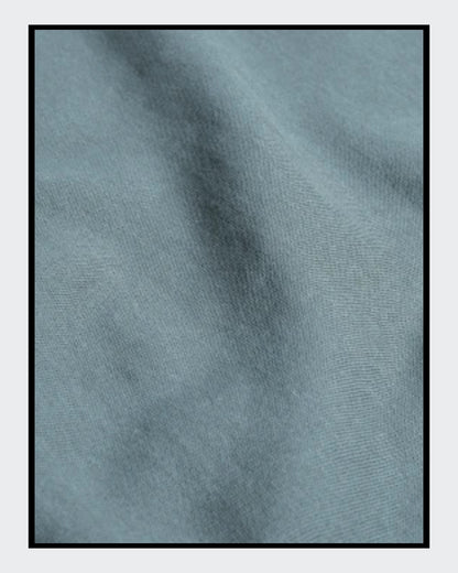 Stoffdetail von: Ein nebelblauer Unisex-Hoodie mit einer Kängurutasche und Kapuze. Auf der Vorderseite ist ein Kunstwerk aufgedruckt: Es zeigt eine stilisierte Frau mit Brille in Collagen-Optik, begleitet von Text in Gelb und Rot. 