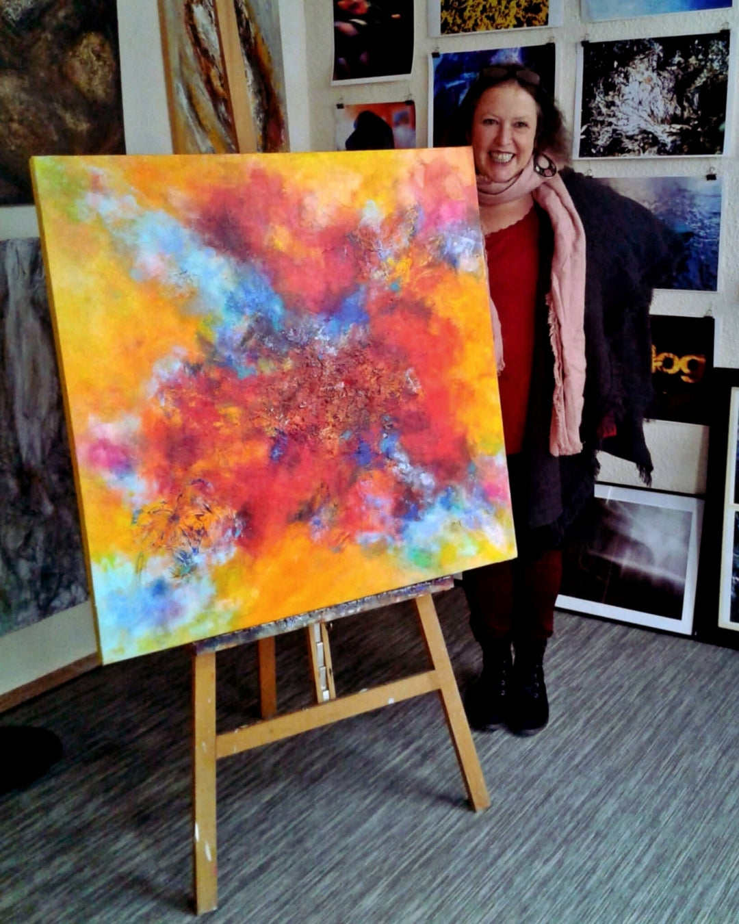 Susanne steht lächelnd in ihrer Galerie. Sie trägt ein rotes Oberteil mit dunkler Jacke, eine dunkle Hose und einen grauen Schal. Hinter ihr steht ein großes, abstraktes Gemälde auf einer Staffelei, dominiert von Gelb-, Rot- und Blautönen. Foto aus der Galerie Paletti aus Wuppertal.