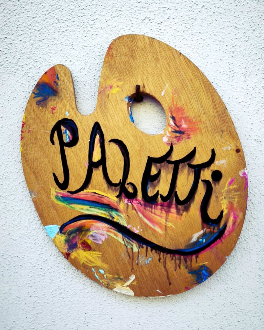 Ein Holzschild in Form einer Malerpalette mit Farbklecksen. In geschwungener schwarzer Schrift steht darauf "Paletti". Das Schild ist an einer weißen Wand befestigt.