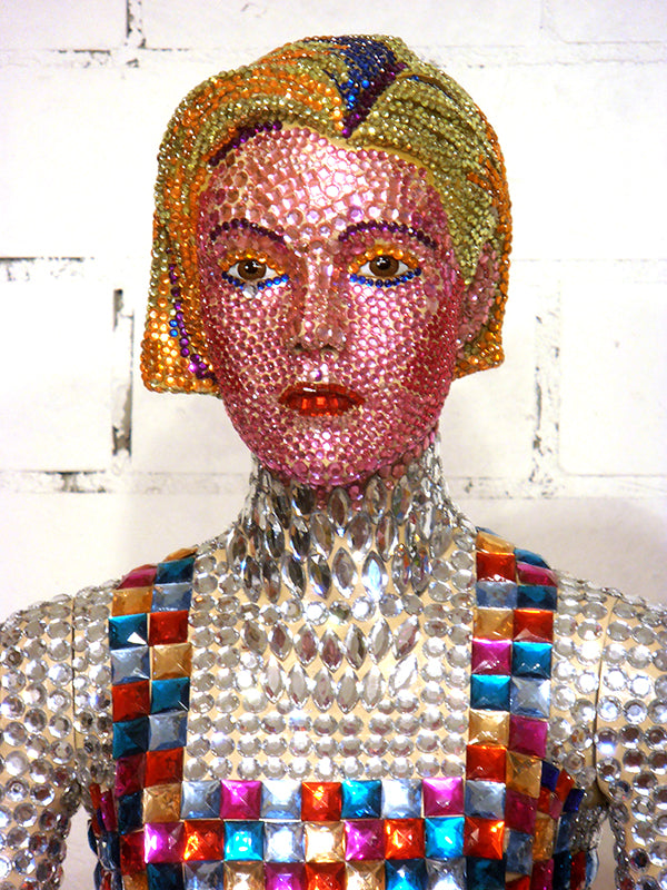 Nahaufnahme einer skulpturalen Puppe, deren Gesicht, Haare und Oberkörper mit vielen bunten, glitzernden Steinen und Elementen besetzt sind. Kunstwerk 