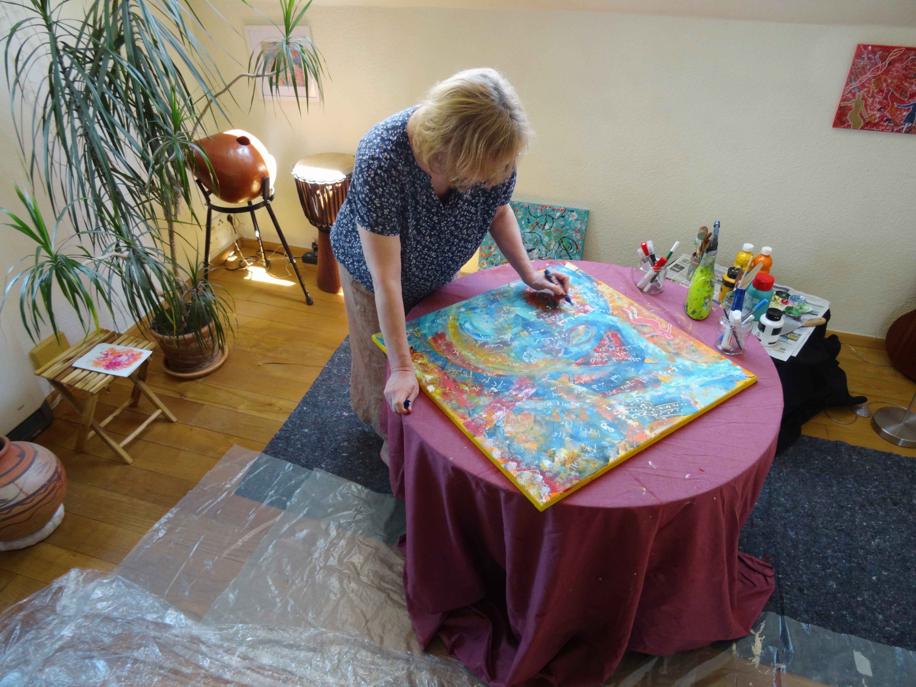 Atelierszene mit einer Künstlerin, die an einem abstrakten, leuchtend bunten Acrylgemälde auf einem runden Tisch arbeitet; umgeben von Pinseln, Farben und inspirierender Studiodekoration.