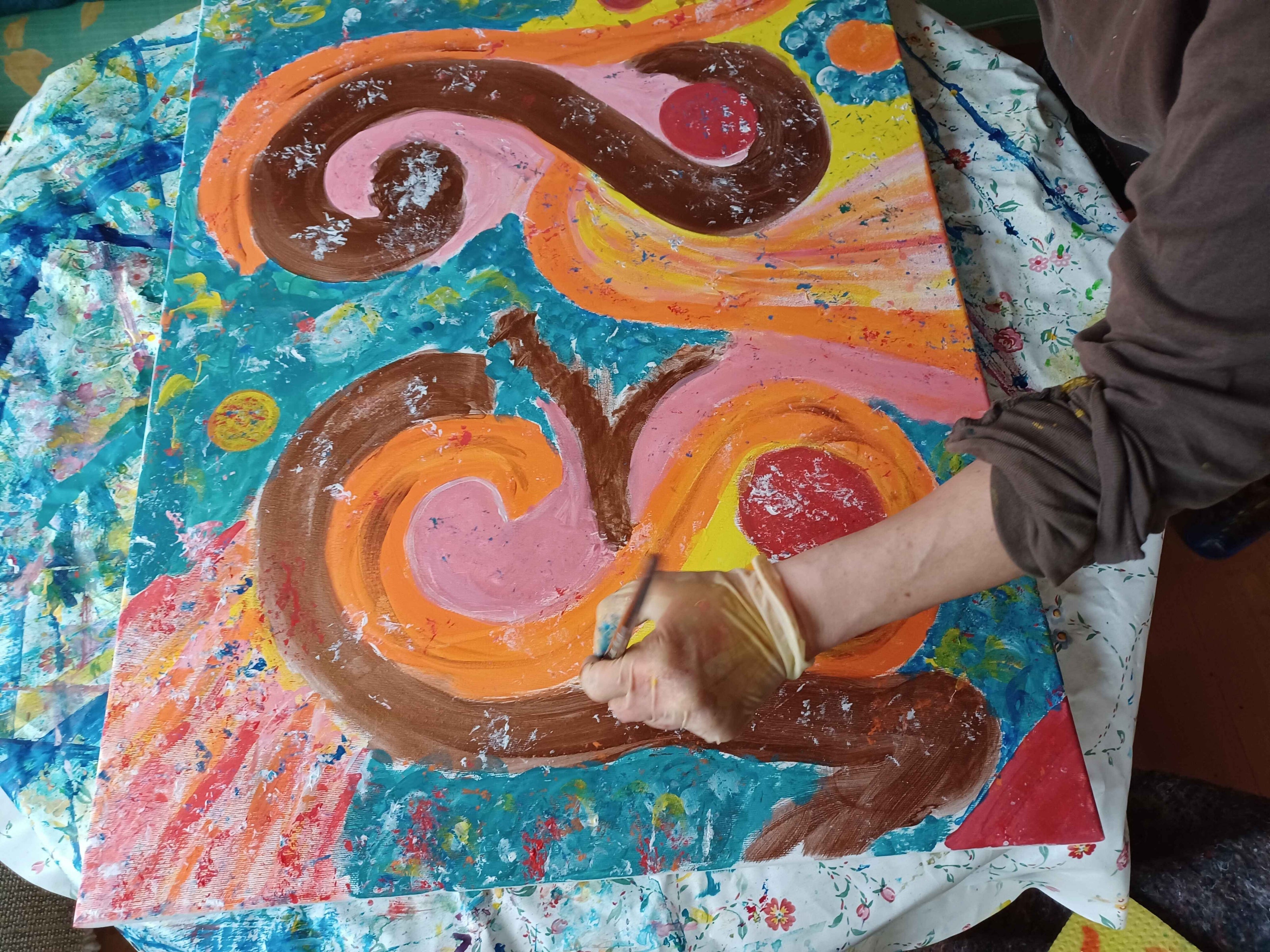 Nahaufnahme des kreativen Arbeitsprozesses: Eine Künstlerin trägt Farbe auf ein abstrahiertes, mehrfarbiges Gemälde auf, das auf einem mit Farbflecken bedeckten Stoff liegt.