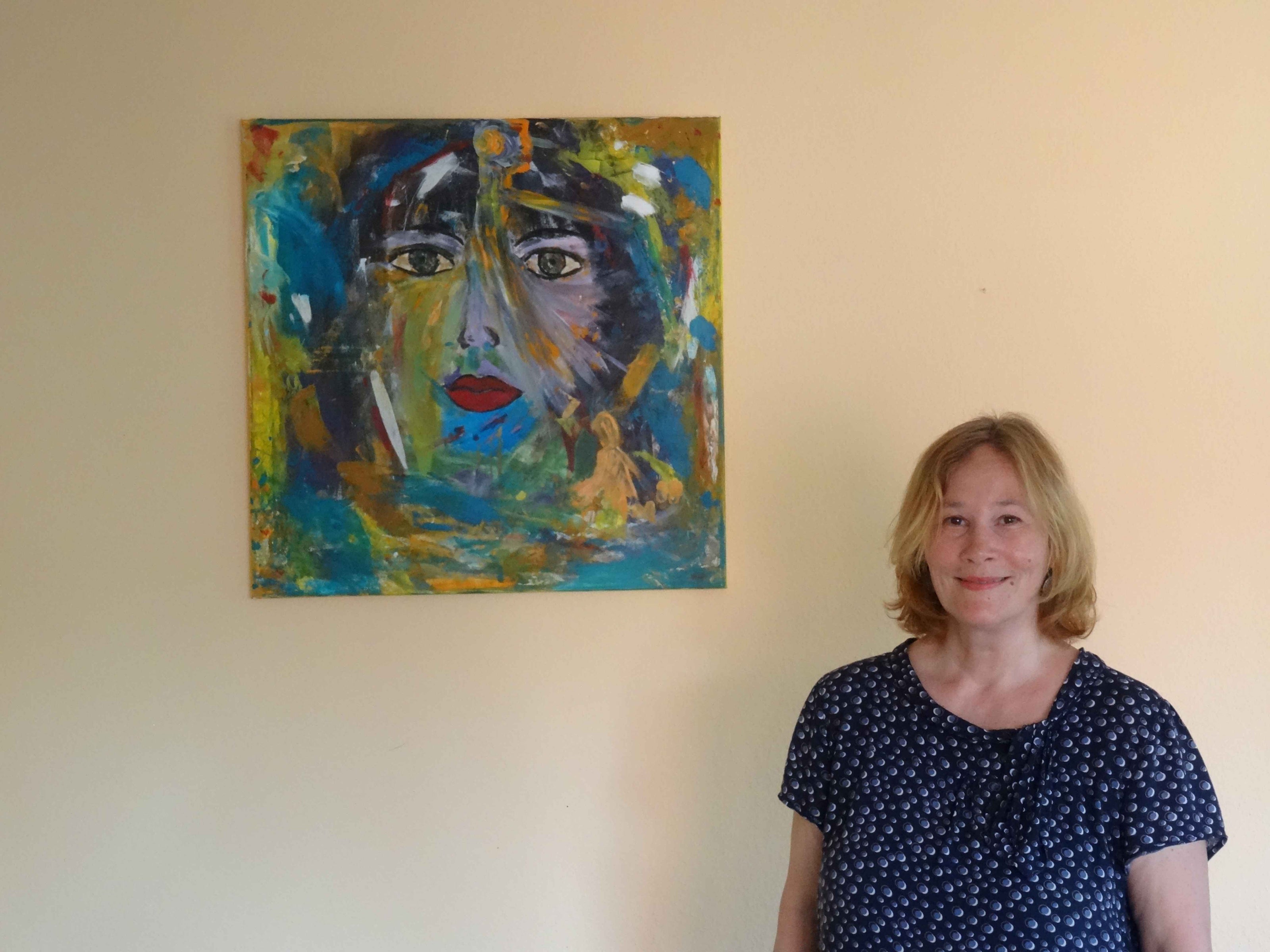 Die Künstlerin steht in einem hellen Atelierraum vor einem farbintensiven abstrakten Gemälde; die ruhige Wandfläche betont die kräftigen Farben und die expressive Malweise des Werkes.