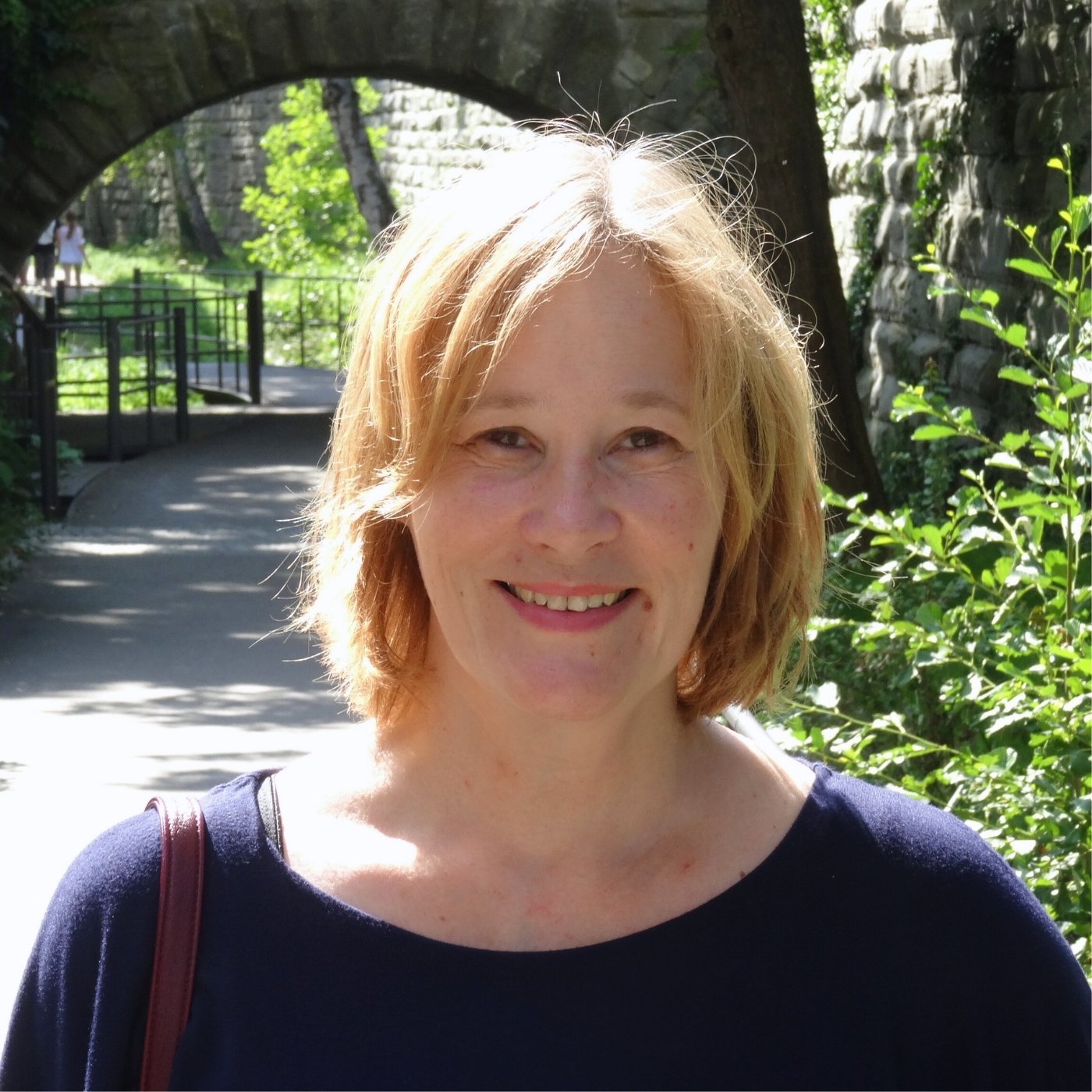 Porträtfoto der Künstlerin Ursula Tüttenberg im Außenbereich, vor einem historischen Steinbogen und von grünem Laub umgeben; sie steht auf einem schattigen Weg in natürlicher, ruhiger Umgebung.