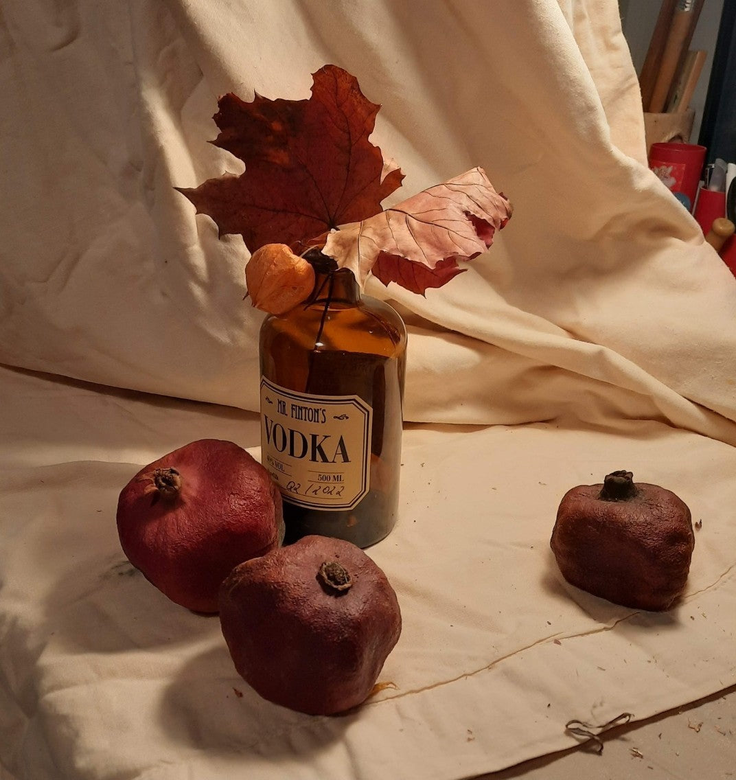 Stillleben-Gemälde "Stillleben mit Granatapfel und Wodka" von Vera Stein. Eine dunkle Wodkaflasche mit Etikett steht auf zerknülltem Papier, umgeben von großen braunen Herbstblättern und drei dunklen, getrockneten Granatäpfeln. Der Hintergrund ist eine Mischung aus Gold, Beige und Braun, teils mit sichtbaren Strukturen. Der Stil ist expressiv und materialreich.