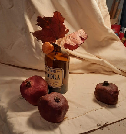 Stillleben-Gemälde "Stillleben mit Granatapfel und Wodka" von Vera Stein. Eine dunkle Wodkaflasche mit Etikett steht auf zerknülltem Papier, umgeben von großen braunen Herbstblättern und drei dunklen, getrockneten Granatäpfeln. Der Hintergrund ist eine Mischung aus Gold, Beige und Braun, teils mit sichtbaren Strukturen. Der Stil ist expressiv und materialreich.
