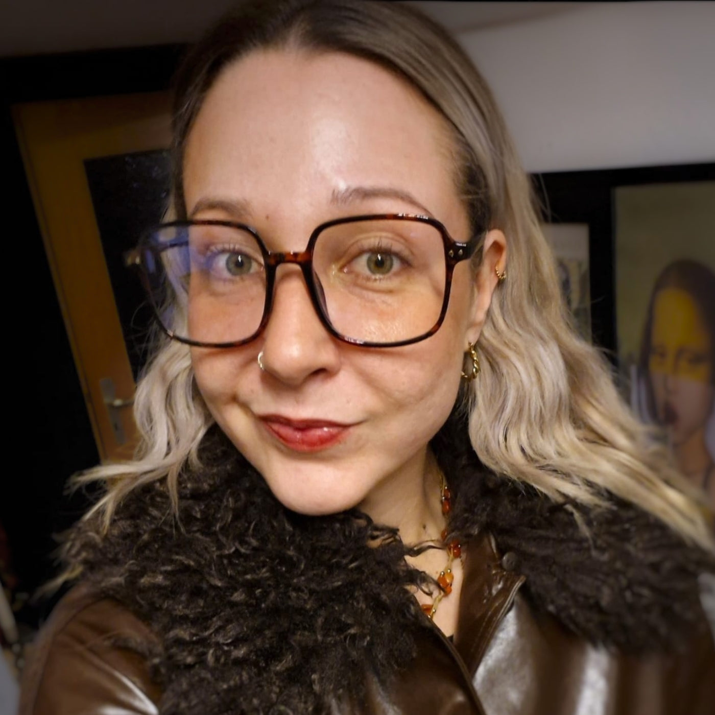 Porträtaufnahme der Künstlerin Yasmin Thiele-Erciyas. Sie blickt mit einem sanften Lächeln direkt in die Kamera und trägt eine markante, quadratische Brille mit dunklem Rahmen sowie eine braune Lederjacke mit dunklem Webpelzkragen. Ihr gewelltes, blondes Haar fällt ihr offen über die Schultern.